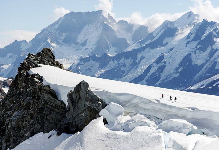 Three people climbing uphill on an ice path towards rocky formations with large mountains in the background