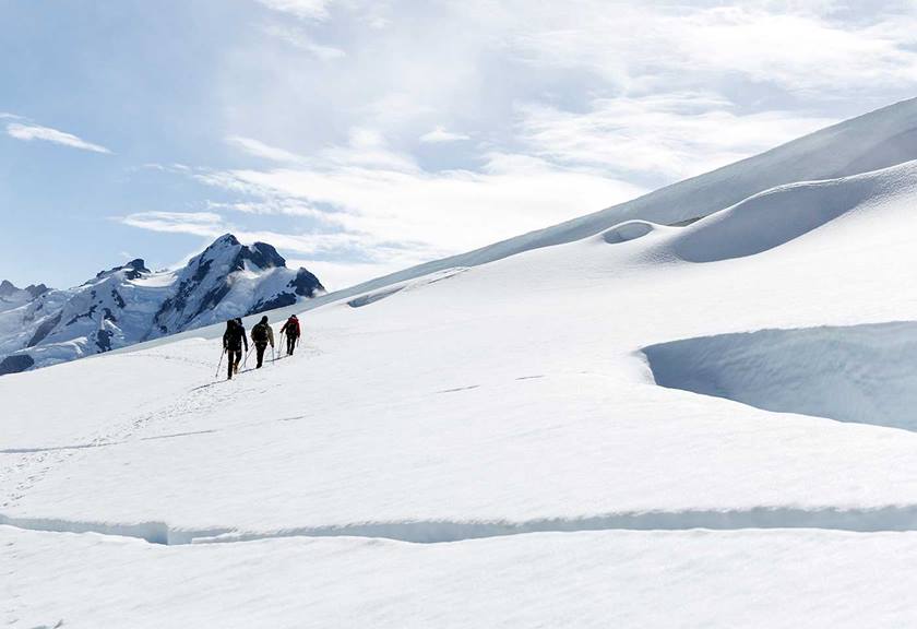 Three people trekking upwards on a snowy slope leaving a trail of footprints