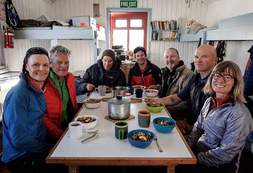 Group of people smiling and sitting around a table with food inside a cabin