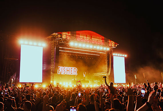 Image of a live show from the back of the crowd showing the full stage with bright screens on the sides