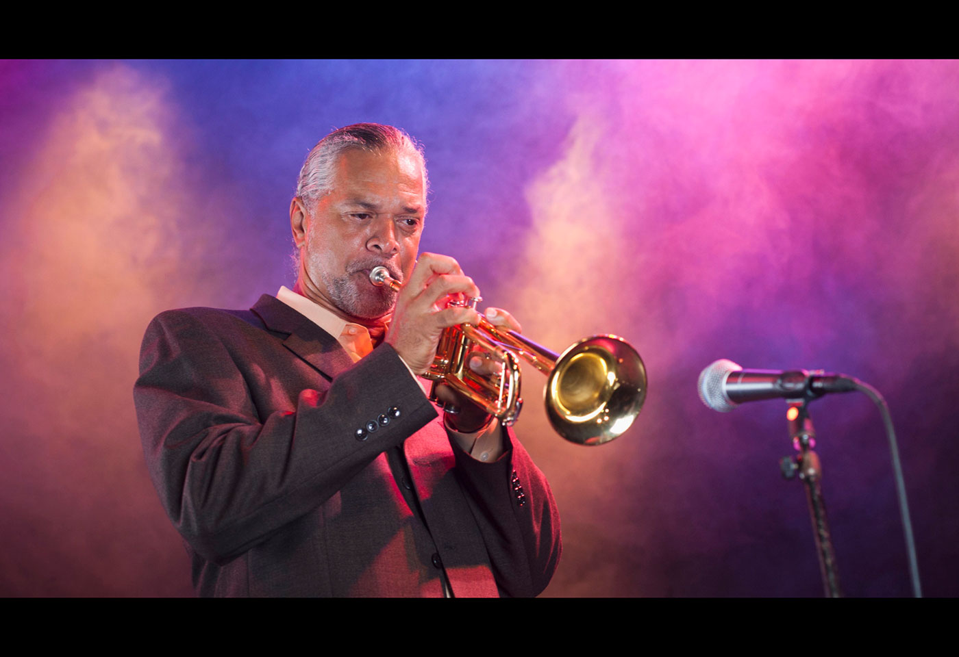 Image of a performer playing the trumpet with a mic in front of him with colourful background