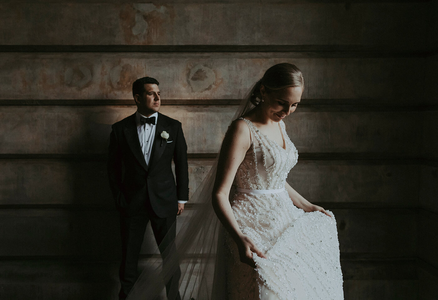 Image of bride looking at her dress with groom on the background by James Simmons